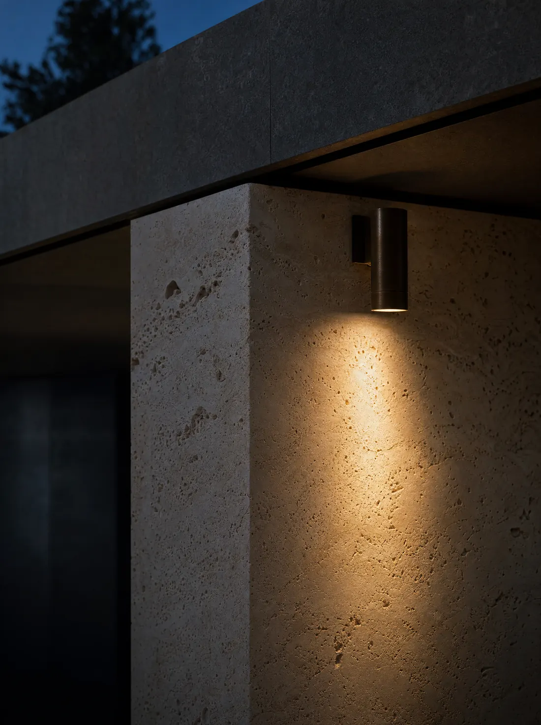 Close-up of a honed travertine pier at dusk, grazed by the beam of a small bronze wall light.